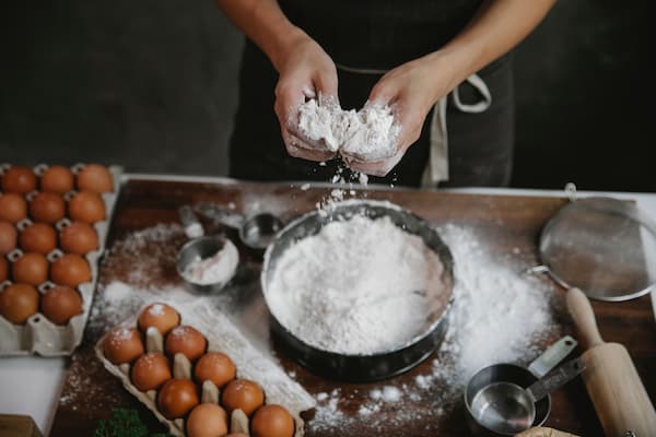 Hands sifting flour into a bowl next to measuring cups, spoons, eggs, and a rolling pin on a wooden baking counter